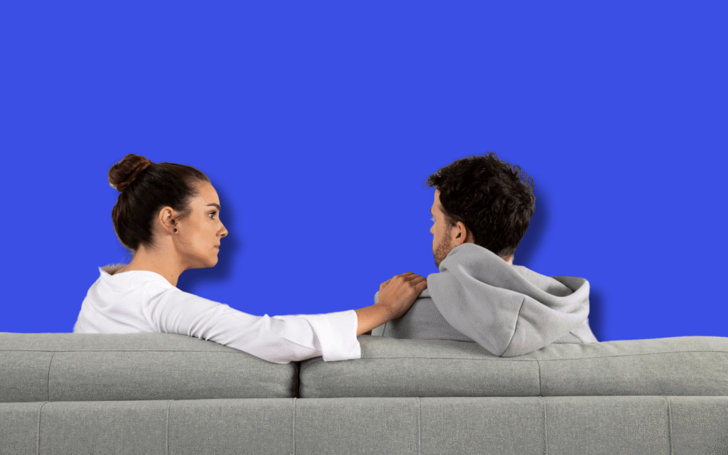 A couple sits on a gray couch holding hands against a blue background, exploring if health insurance covers couples therapy. Find out how coverage works, with many plans now including therapy to support relationships.