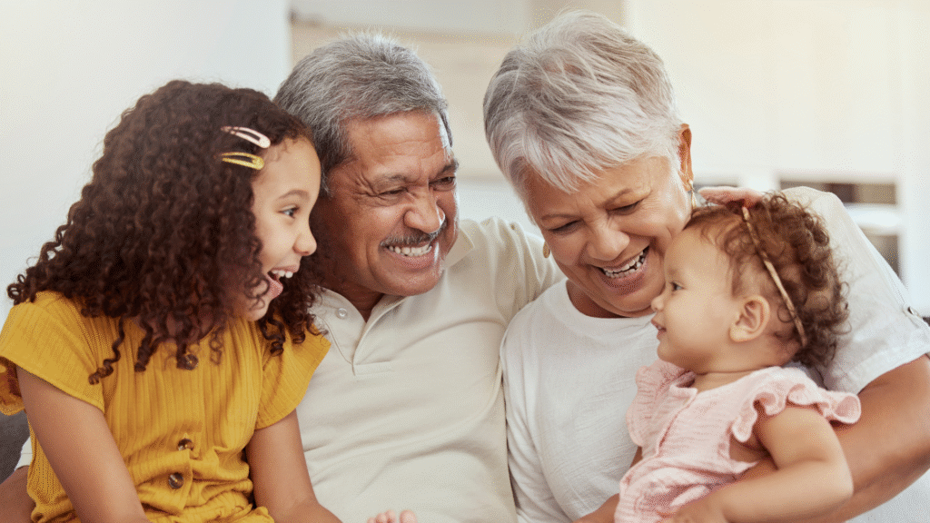 A joyful family scene with grandparents and grandchildren, featuring a smiling elderly couple holding a baby and a young girl in a yellow dress, all sharing a warm moment.