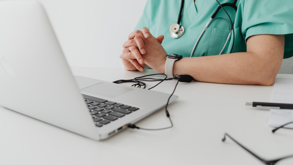 Image of a doctor in green scrubs with a stethoscope, sitting at a desk with clasped hands, a laptop, smartwatch, and glasses, symbolizing healthcare and technology integration.